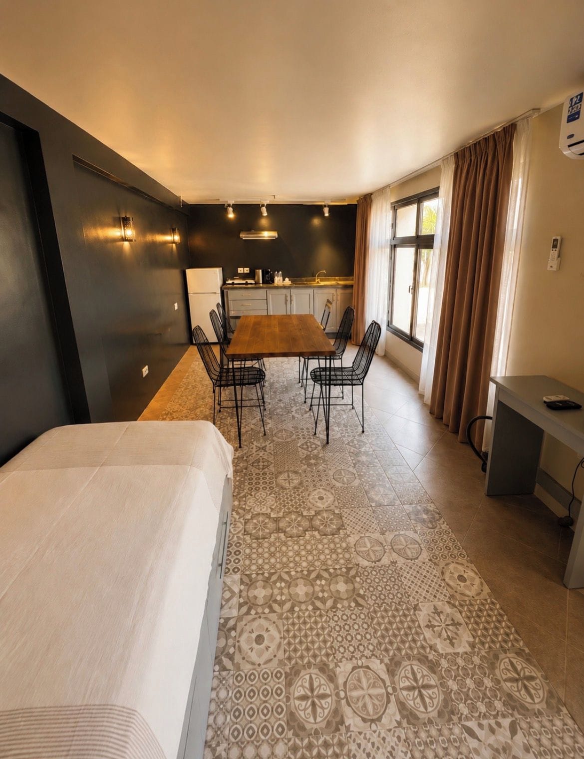 A modern hotel room with a bed, a dining table with chairs, a kitchen area, and a small desk.