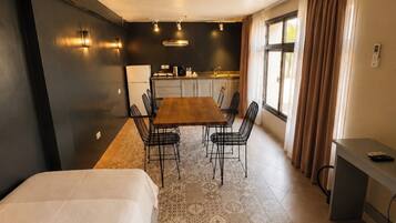 A modern hotel room with a bed, a dining table with chairs, a kitchen area, and a small desk.