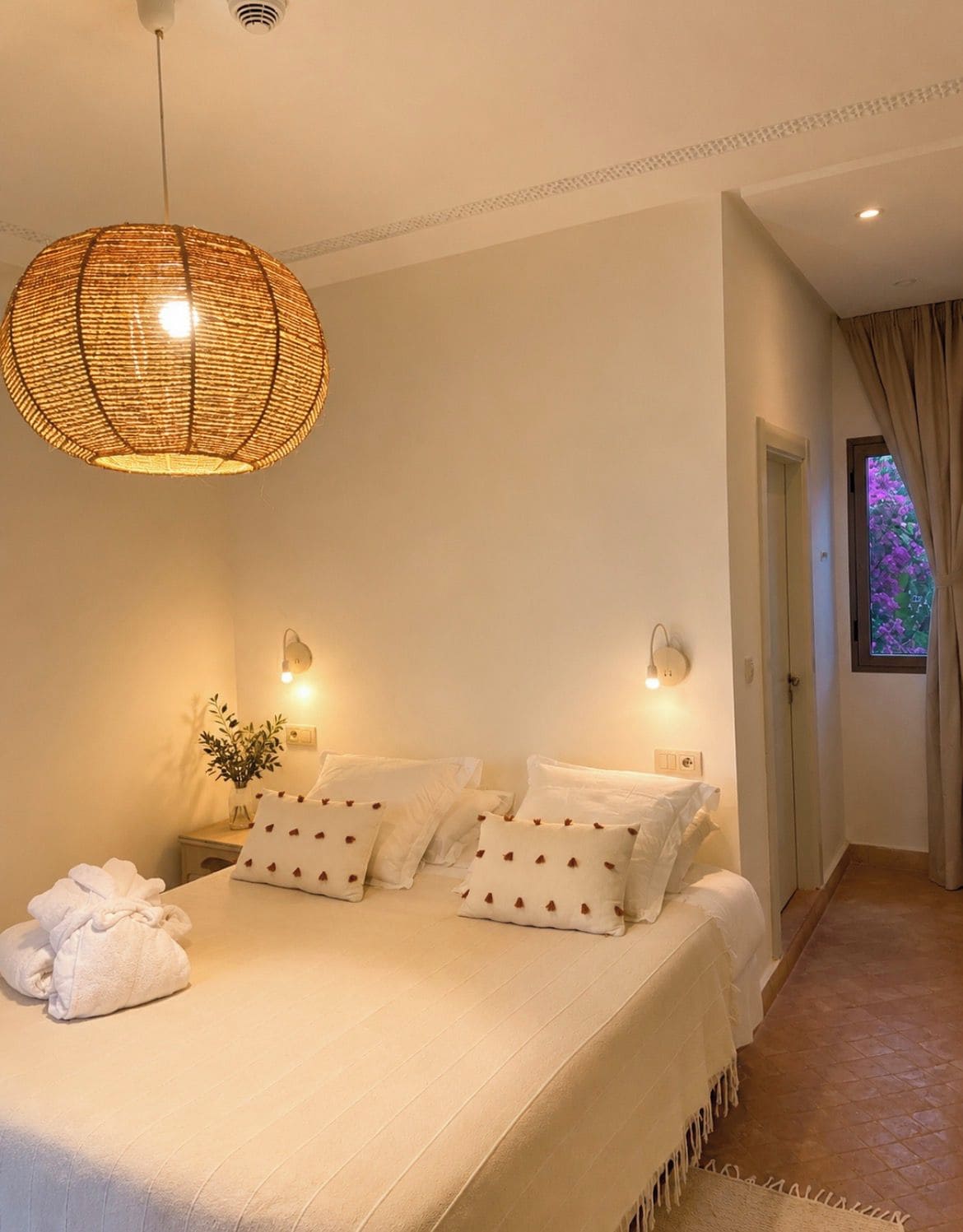 Un dormitorio con una cama grande, lámparas de noche, una lámpara colgante y vistas de un jardín a través de una ventana.