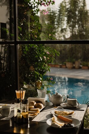 Une table de petit-déjeuner avec du thé, du café et des viennoiseries, placée près d’une fenêtre donnant sur une piscine.