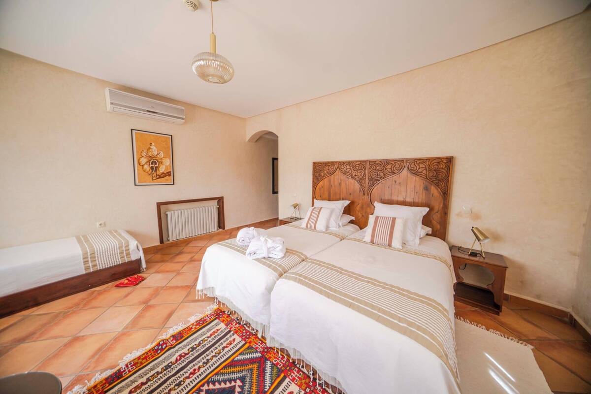 A hotel room with a large bed, a smaller bed, a wooden headboard, a nightstand, a lamp, a framed picture on the wall, and a patterned rug.