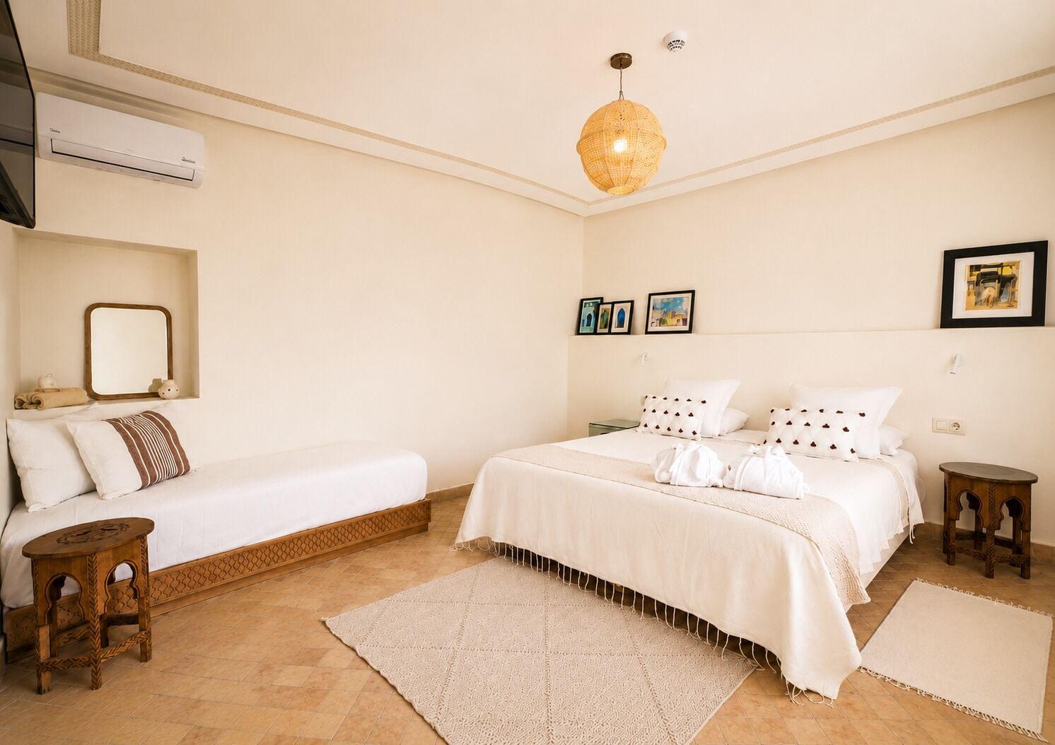 Habitación de hotel con cama, mesita de noche, taburete de madera, alfombra grande tejida y lámpara en el techo.