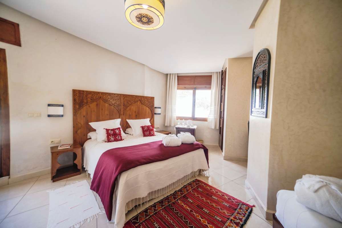 Une chambre à coucher avec une tête de lit en bois, un grand lit avec une literie blanche et rouge, un tapis à motifs rouges et blancs, une table de chevet avec une lampe et une fenêtre avec des rideaux.