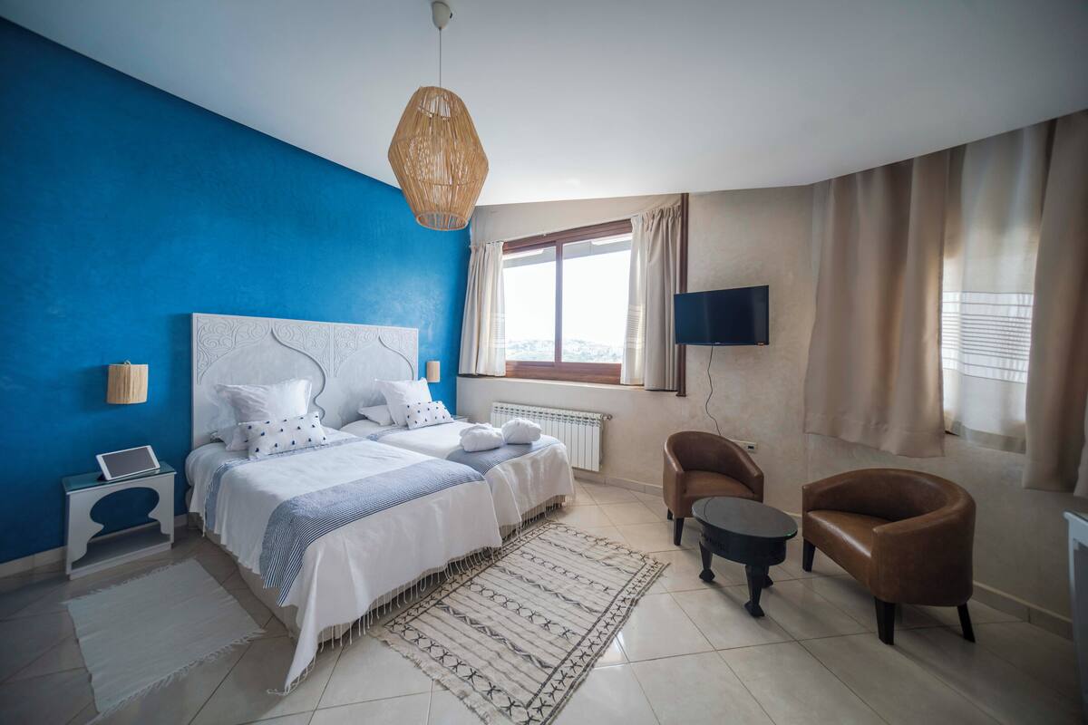 A bedroom with a large bed, a TV mounted on the wall, a window with curtains, and a chair.