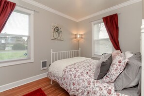 A bedroom with a white metal bed frame, floral bedding, and red curtains. There is a wall-mounted lamp and a window with blinds.
