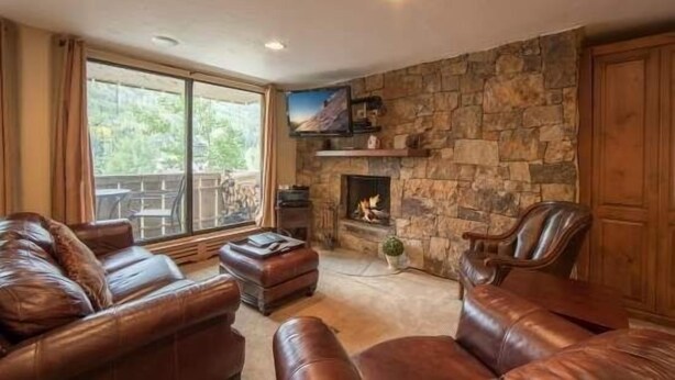 A cozy living room with leather sofas, a stone fireplace, and a large window overlooking a deck. - Scorpio Condos with Great Vail Mountain Views by RedAwning (Vail)
