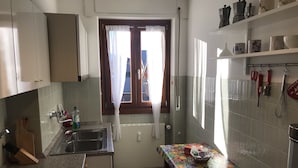 Eine kleine Küche mit Spüle, einem Fenster mit Vorhängen, einem Tisch mit bunten Tischdecken und verschiedenen Küchenutensilien an der Wand.
