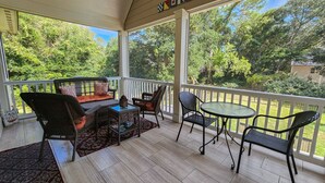 Une véranda couverte, meublée de meubles en osier, avec une table en verre et une vue sur la verdure environnante.