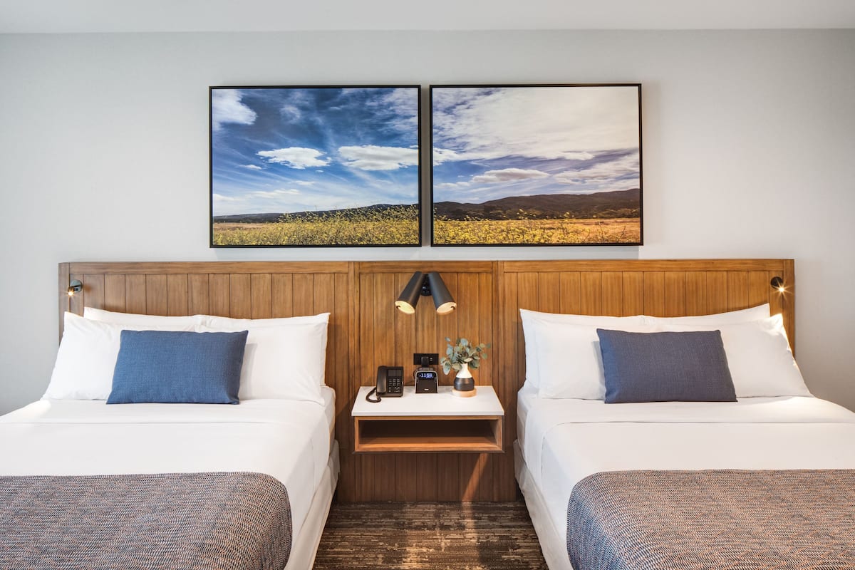 Habitación de hotel con dos camas, cabecero de madera, mesita de noche con lámpara, teléfono y un jarrón con flores.