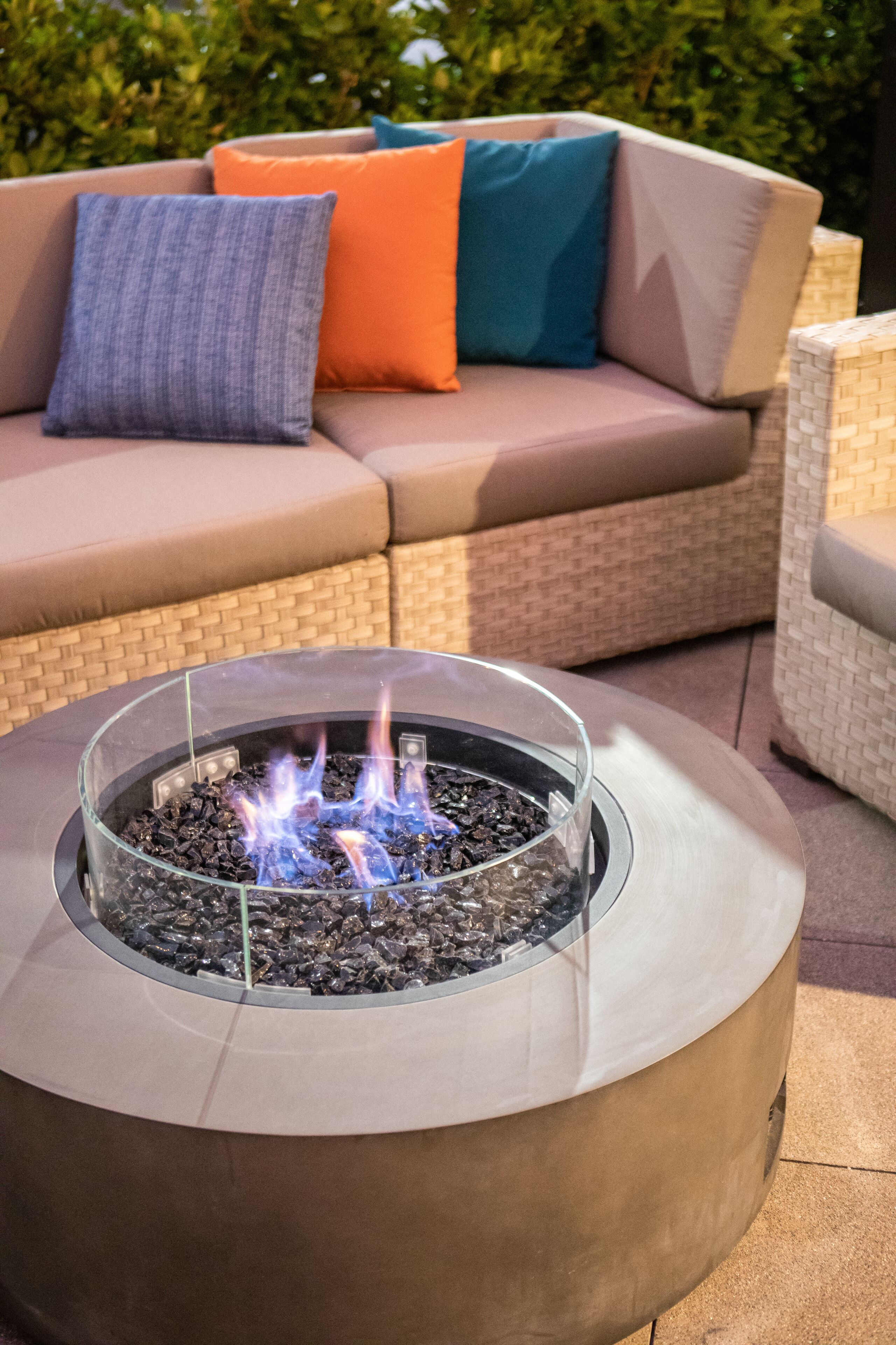 A round outdoor fire pit with visible flames, surrounded by a beige sofa with orange and blue cushions, and wicker furniture in the background.