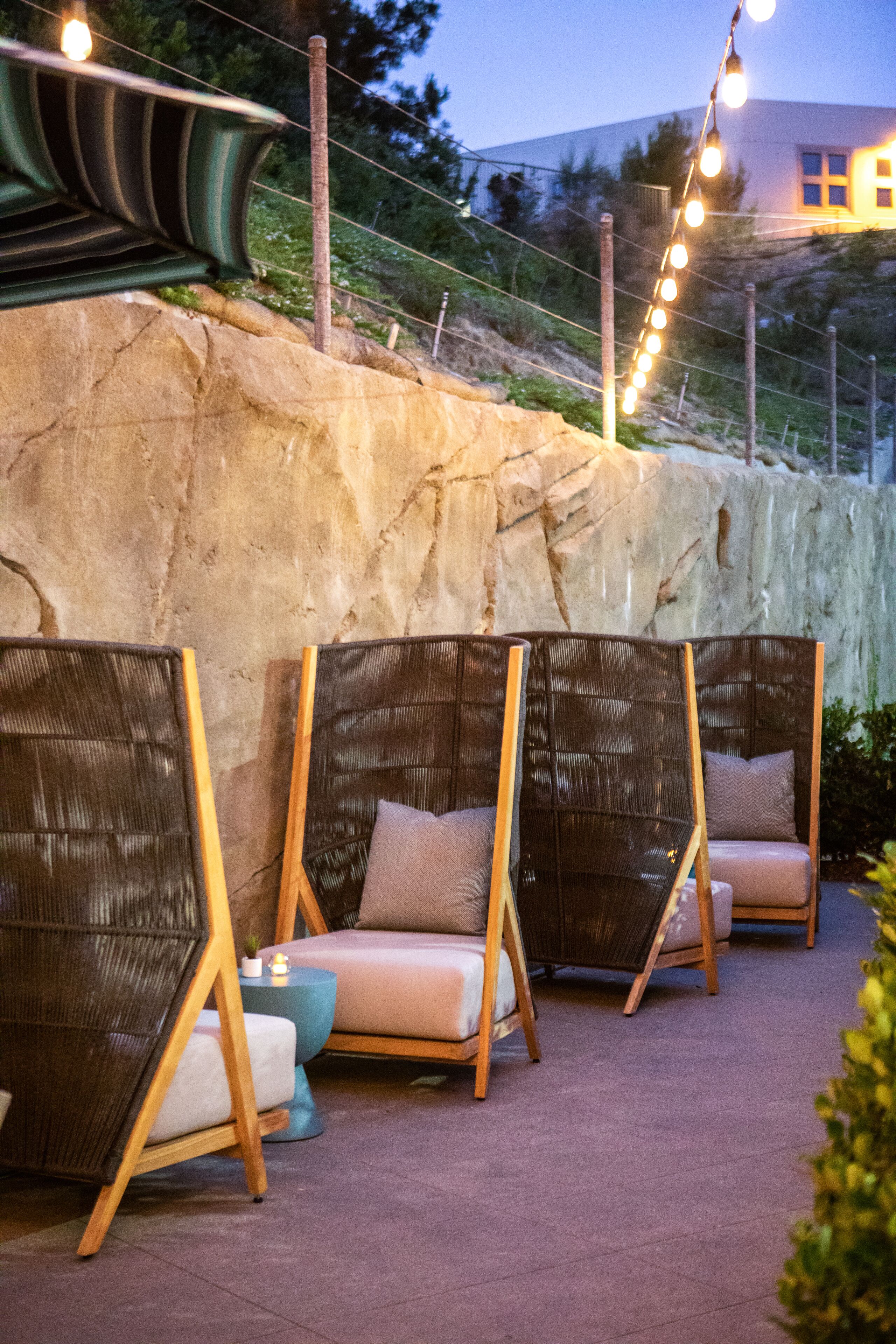 Outdoor seating with wicker chairs and cushions, a stone wall, and string lights.
