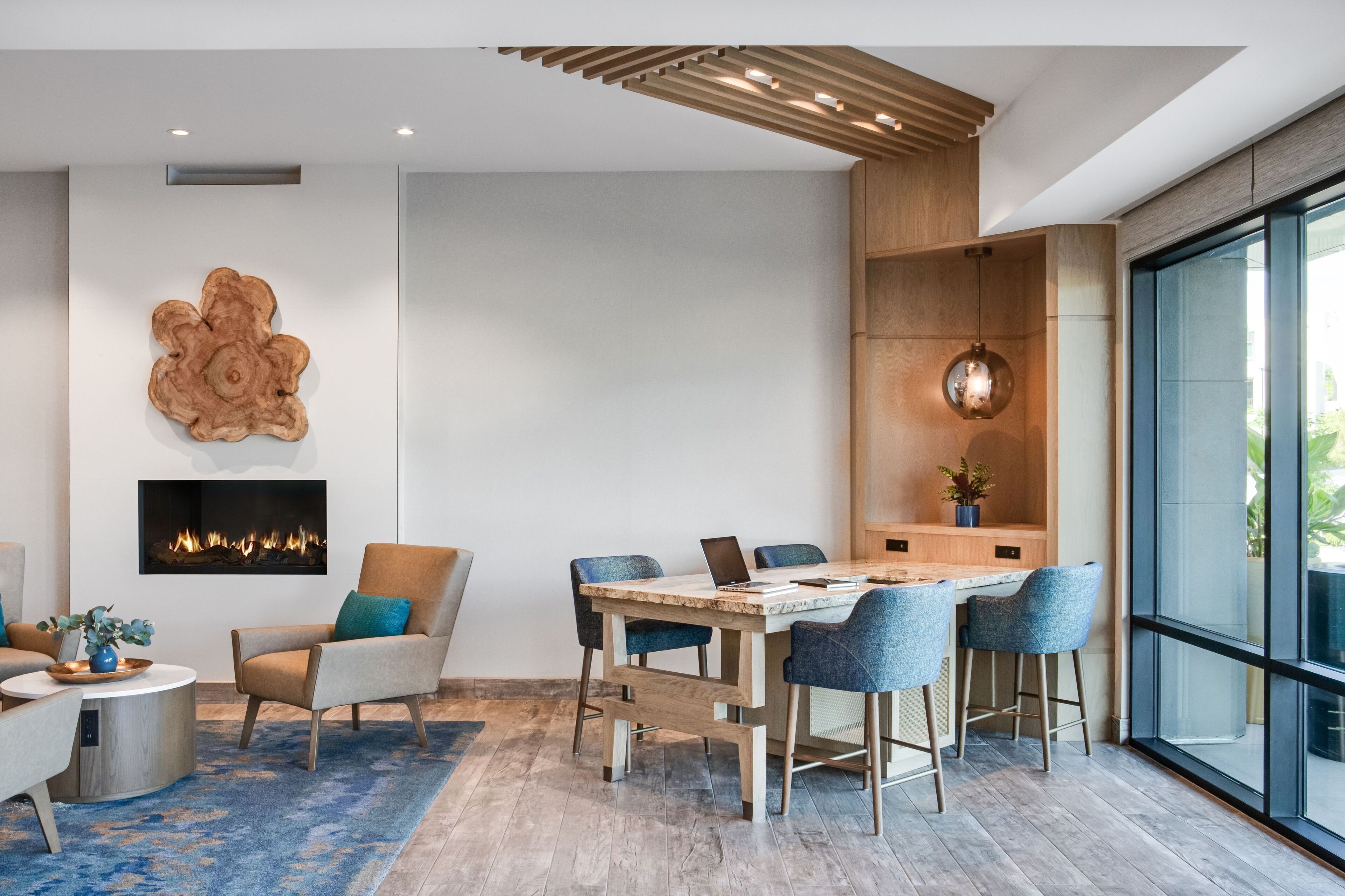 A modern living room with a fireplace, a dining area with a wooden table and chairs, and a large window offering a view of the outdoors.