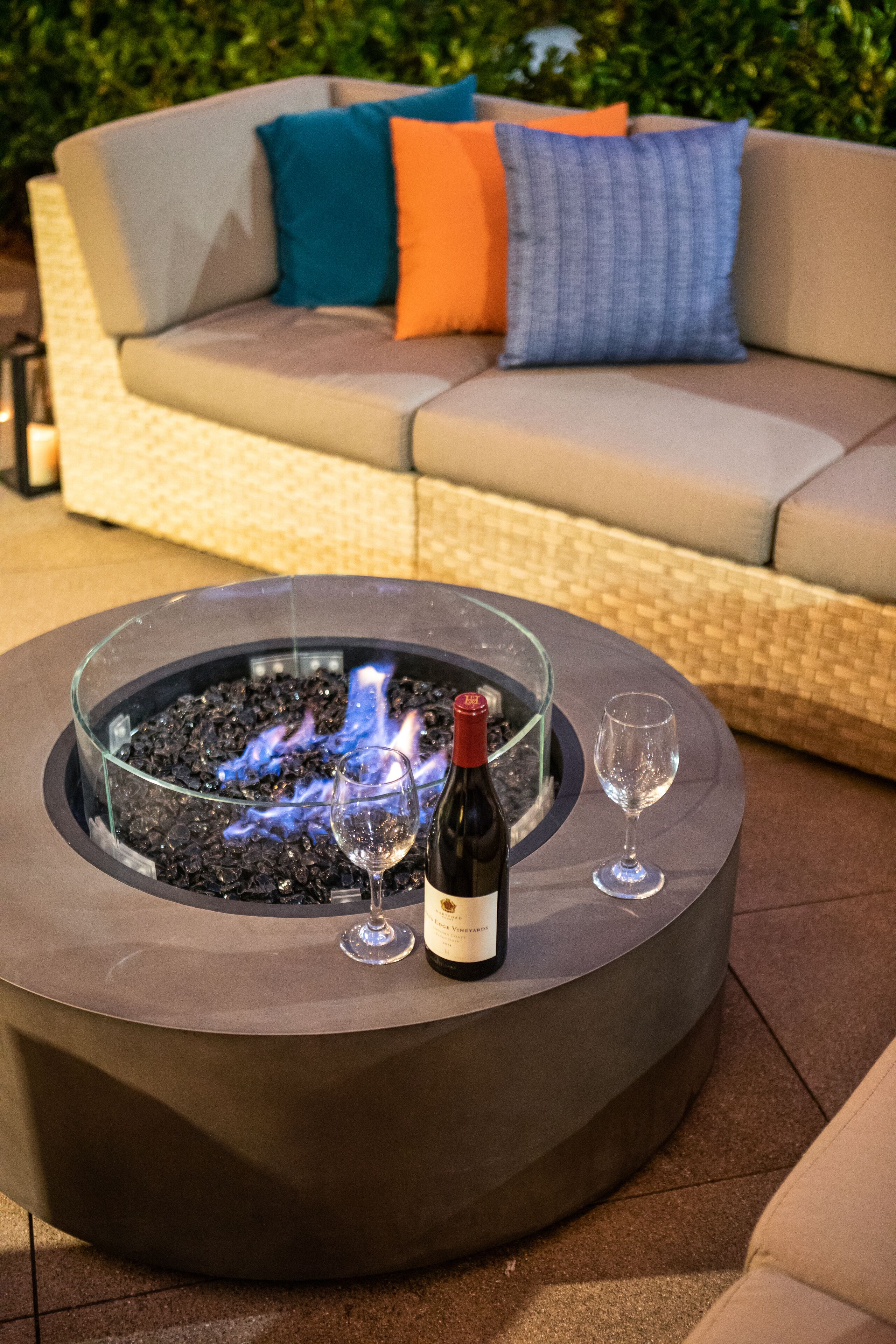 Fuego exterior circular con recinto de vidrio, dos copas de vino y una botella de vino en un patio.