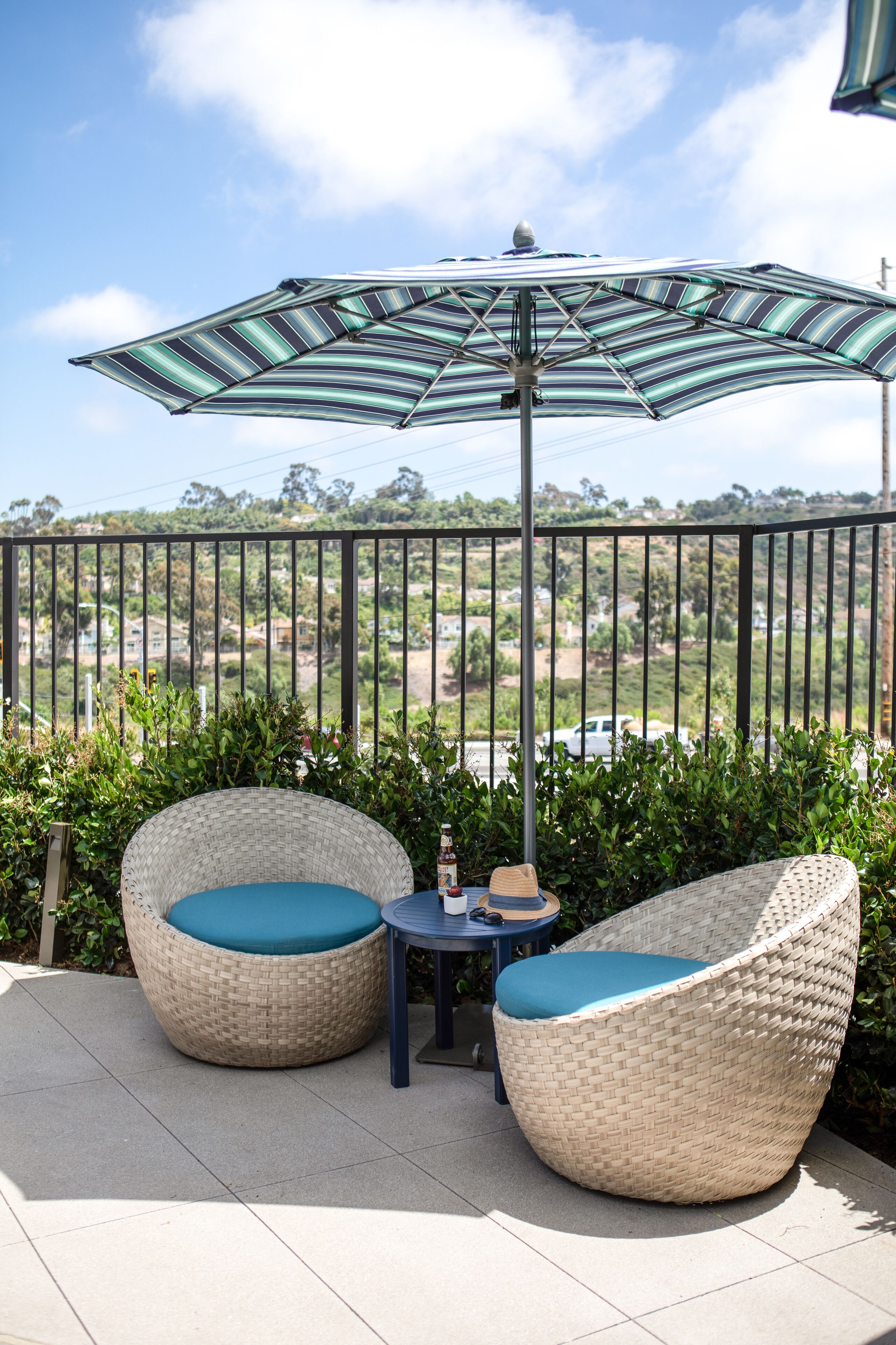 A balcony with wicker chairs and a table, a striped umbrella, and a view of a residential area.