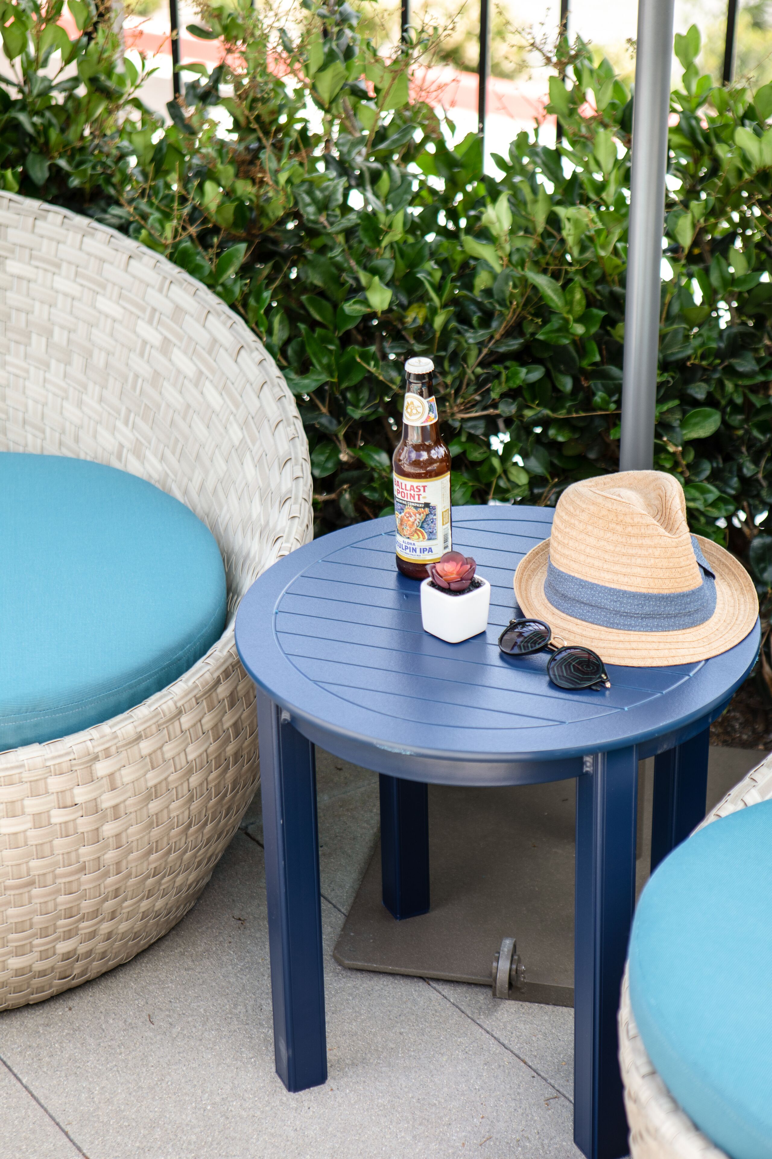 A wicker chair with a blue cushion, a small round table with a bottle of beer, a hat, and sunglasses.