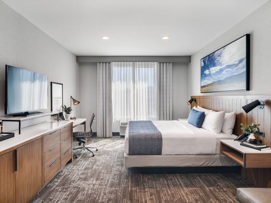 A modern hotel room with a large bed, a desk with a flat-screen TV, a coffee maker, and a large window with curtains.