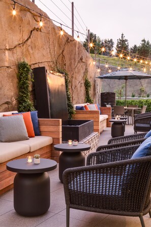 Un área de estar al aire libre con sofás, sillones y mesas, rodeada por un muro de piedra y luces colgantes.