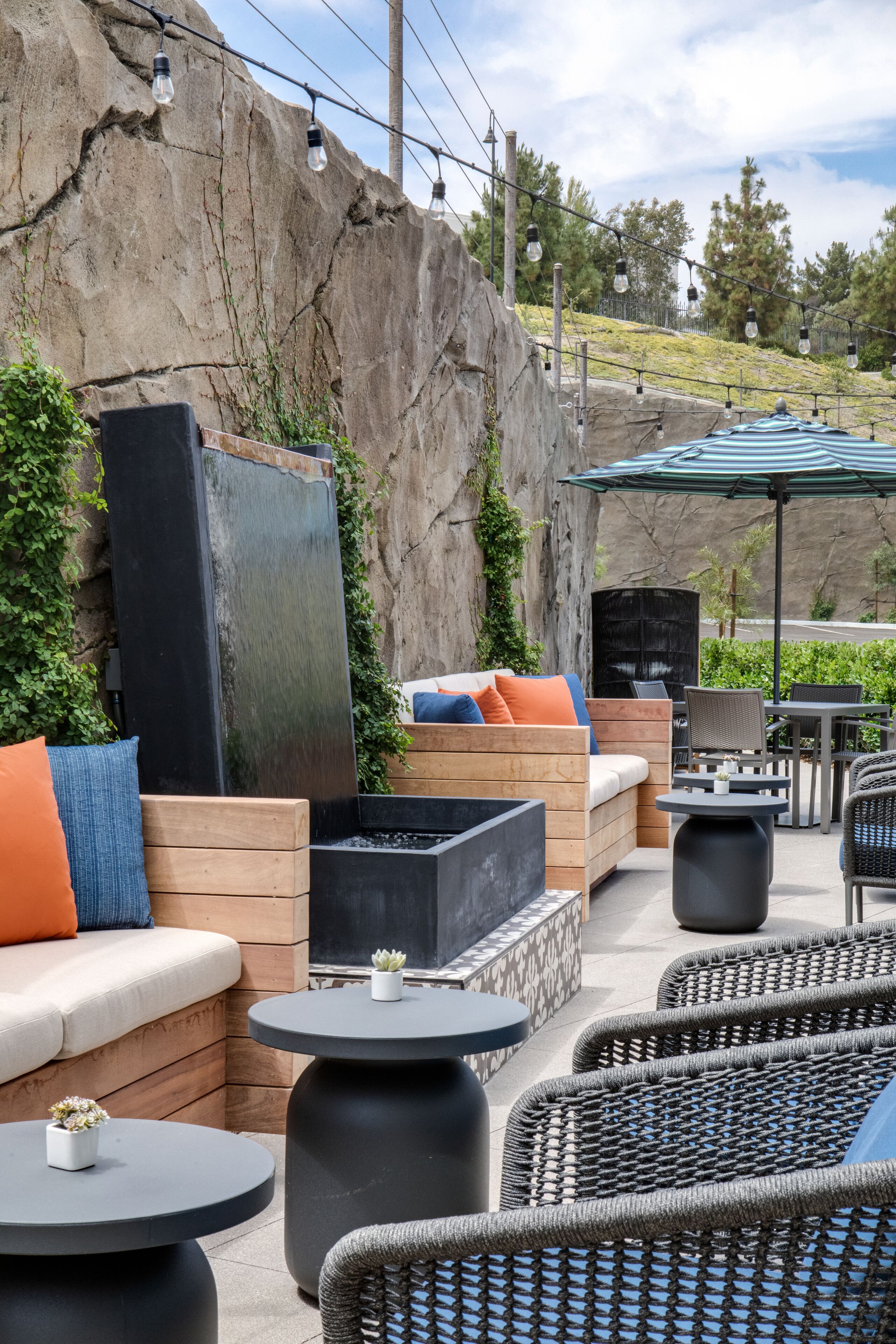 An outdoor seating area with wooden benches, round tables, and a large rock wall.