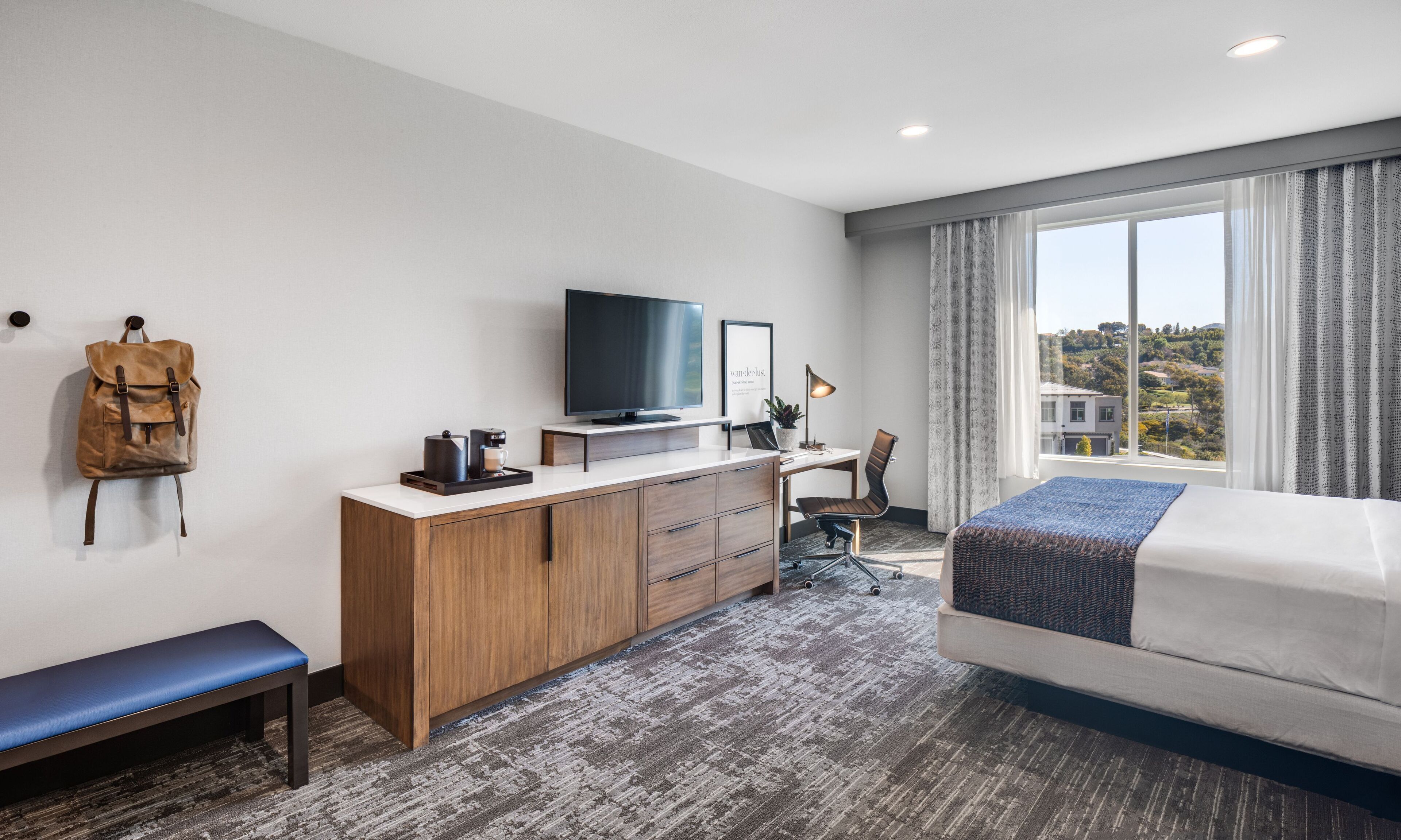 A hotel room with a bed, a desk with a TV, a chair, a bench, and a backpack on the wall.