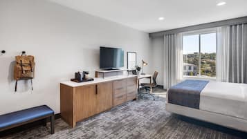 A hotel room with a bed, a desk with a TV, a chair, a bench, and a backpack on the wall.