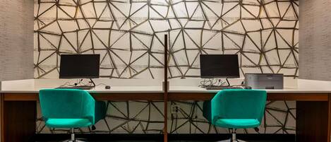 A conference room with two desks, each with a computer and a printer. The room has a geometric patterned wall and carpeted floor.