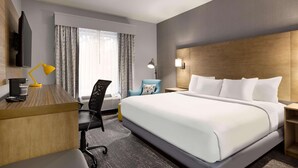 A hotel room with a large bed, a desk with a lamp, a chair, a TV, and a window with curtains.