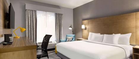 A hotel room with a large bed, a desk with a lamp, a chair, a TV, and a window with curtains.