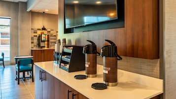 A modern hotel bar area with a flat-screen TV mounted on the wall, a coffee machine, and a seating area with chairs and a table.