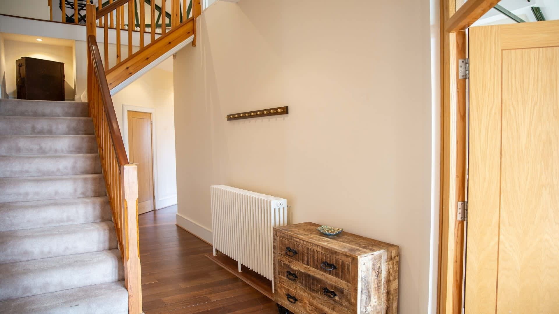 A staircase with wooden railings leading to a landing. A white radiator is mounted on the wall, and a rustic wooden chest of drawers is placed against the wall.