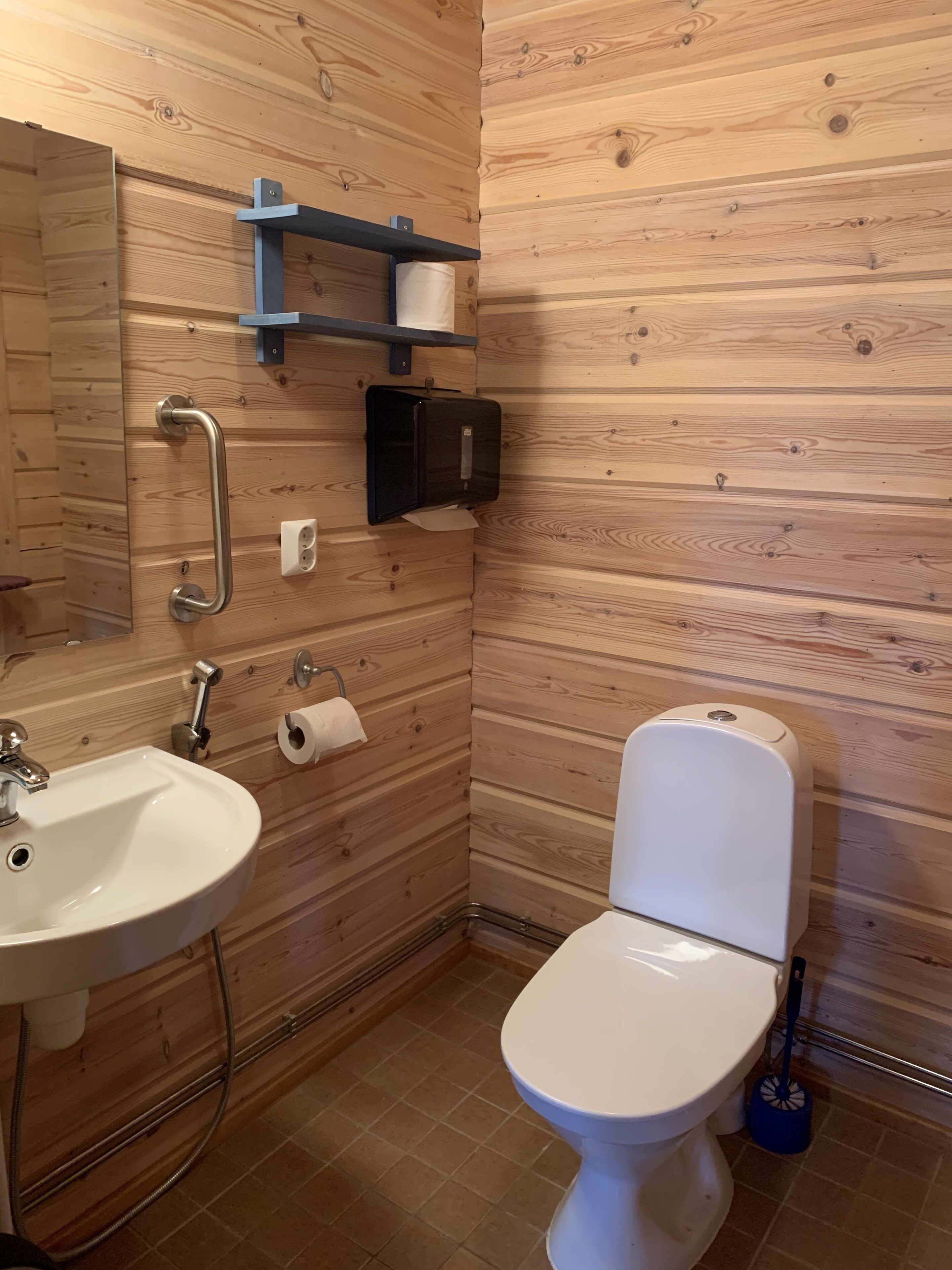 Un baño con pared de madera, un lavamanos blanco, un inodoro, un espejo y una estantería con una toalla.