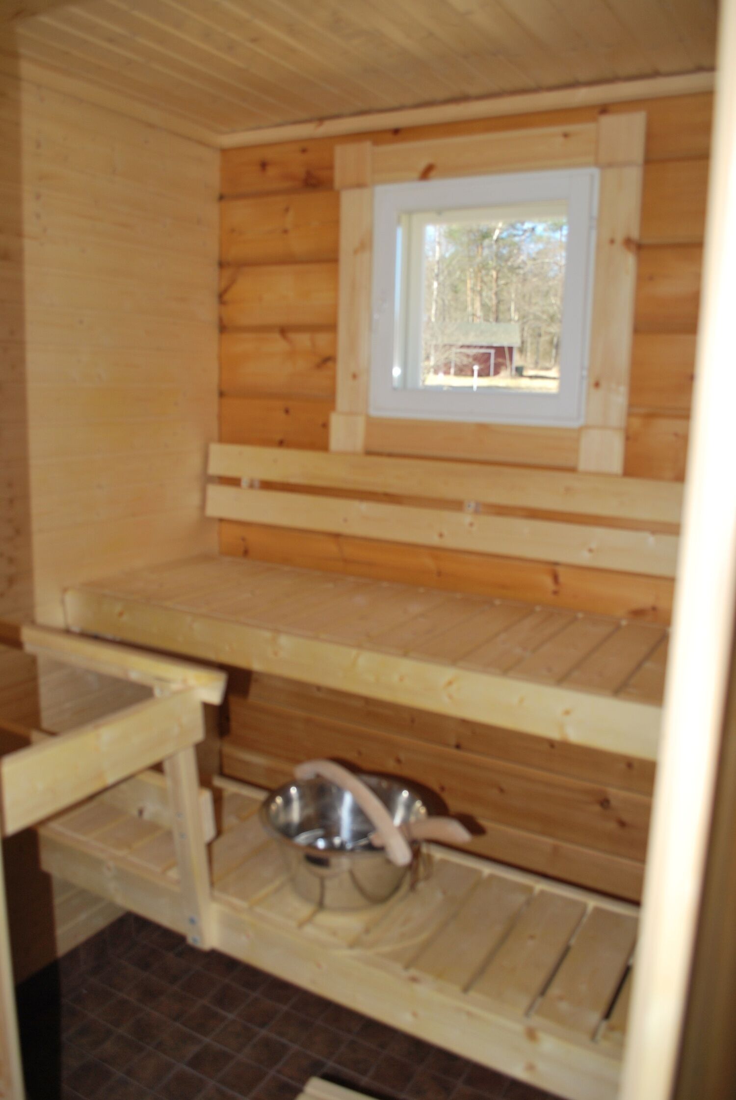 Interior de una cabaña de madera con una ventana, un recipiente metálico y un banco de madera.