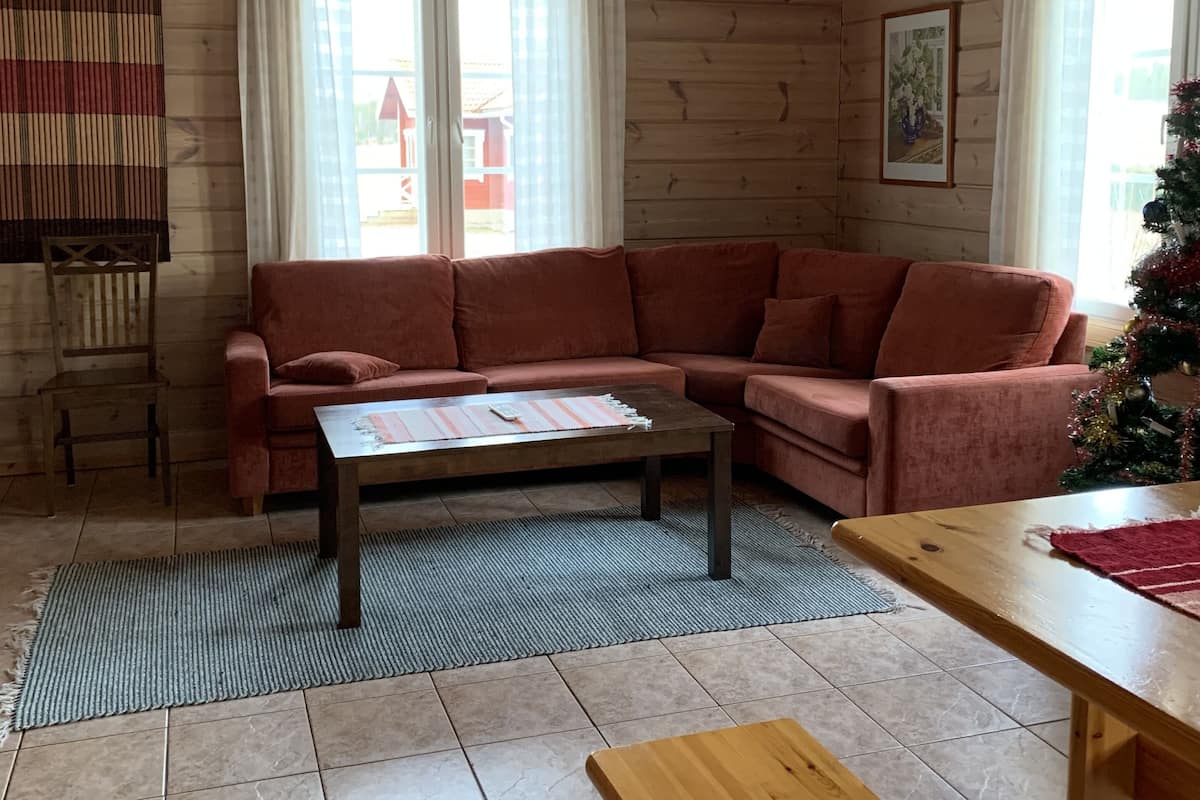 A cozy living room with a wooden ceiling, a sectional sofa, a coffee table, a wooden bench, and a large window with curtains.