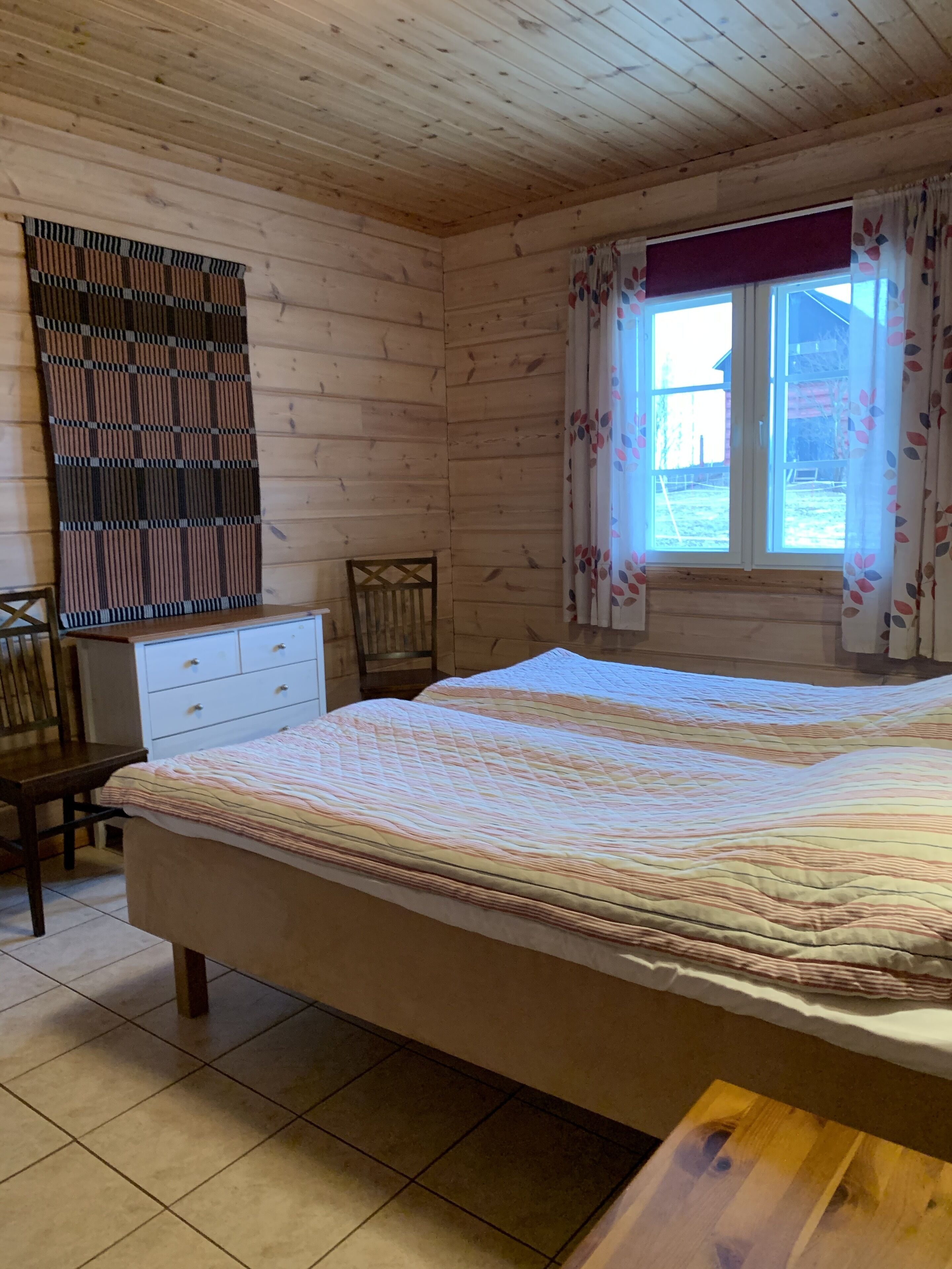 Un dormitorio con paneles de madera, una cama, un tocador, una silla y una ventana con cortinas florales.