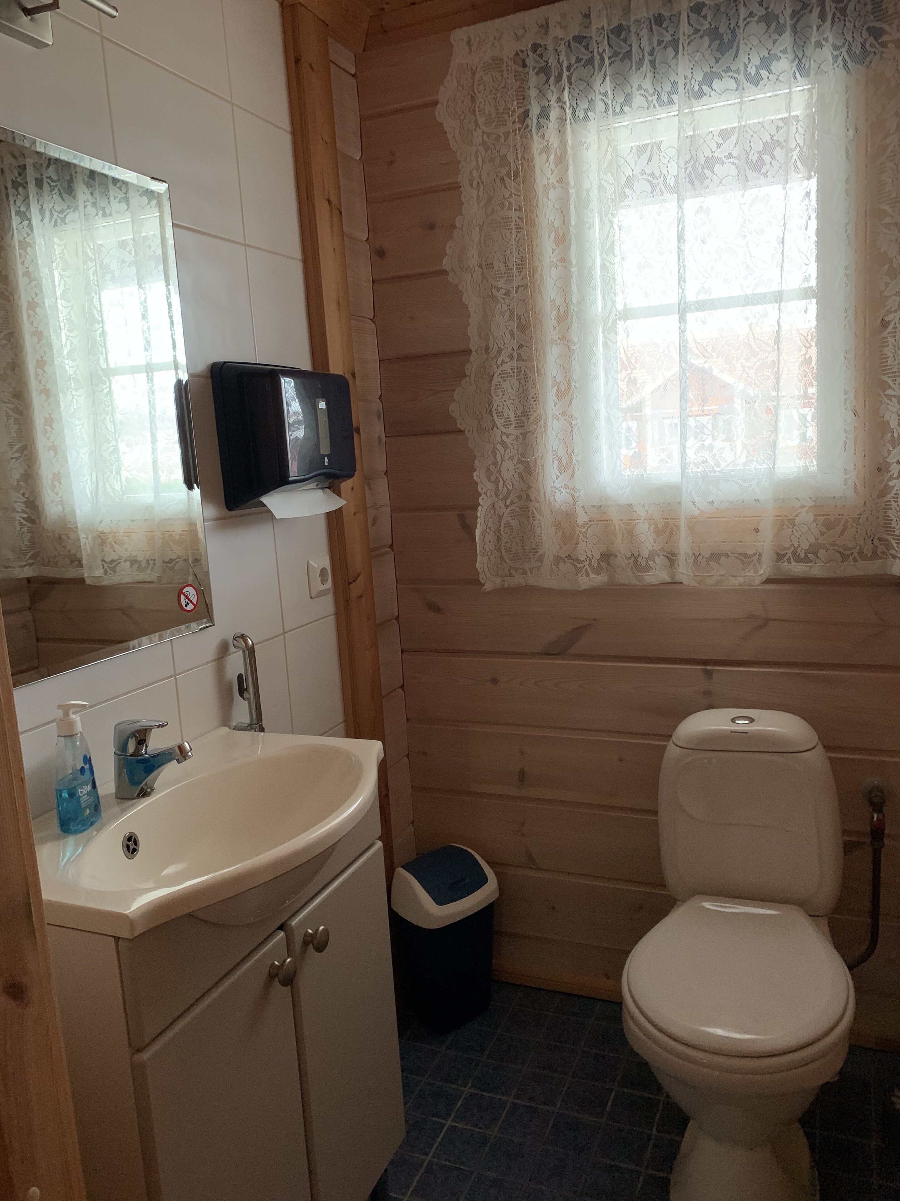 Un baño con lavamanos, espejo, inodoro y ventana con cortinas de encaje.