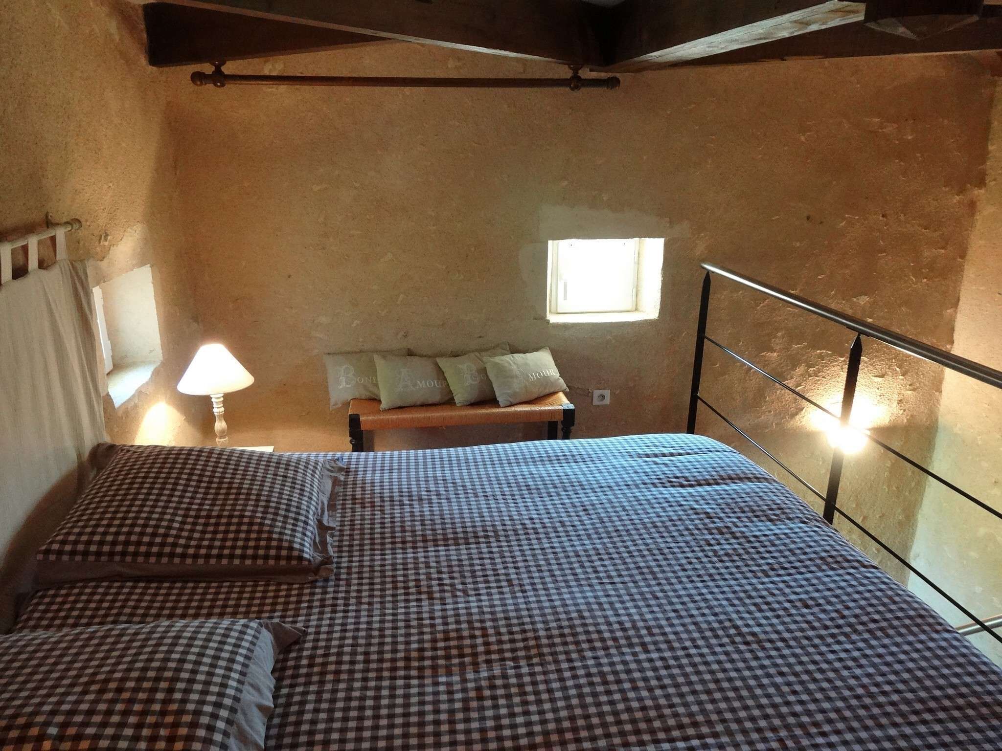 A bedroom with a checkered bedspread, a nightstand with a lamp, a window with a view, and a wooden bench with pillows.