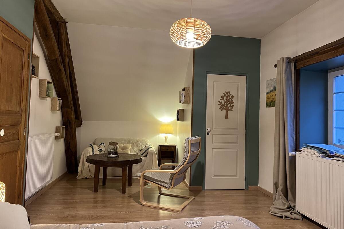 Une pièce chaleureuse avec une table ronde, un fauteuil confortable, un luminaire suspendu, une porte décorée d’un motif d’arbre et une fenêtre avec des rideaux.