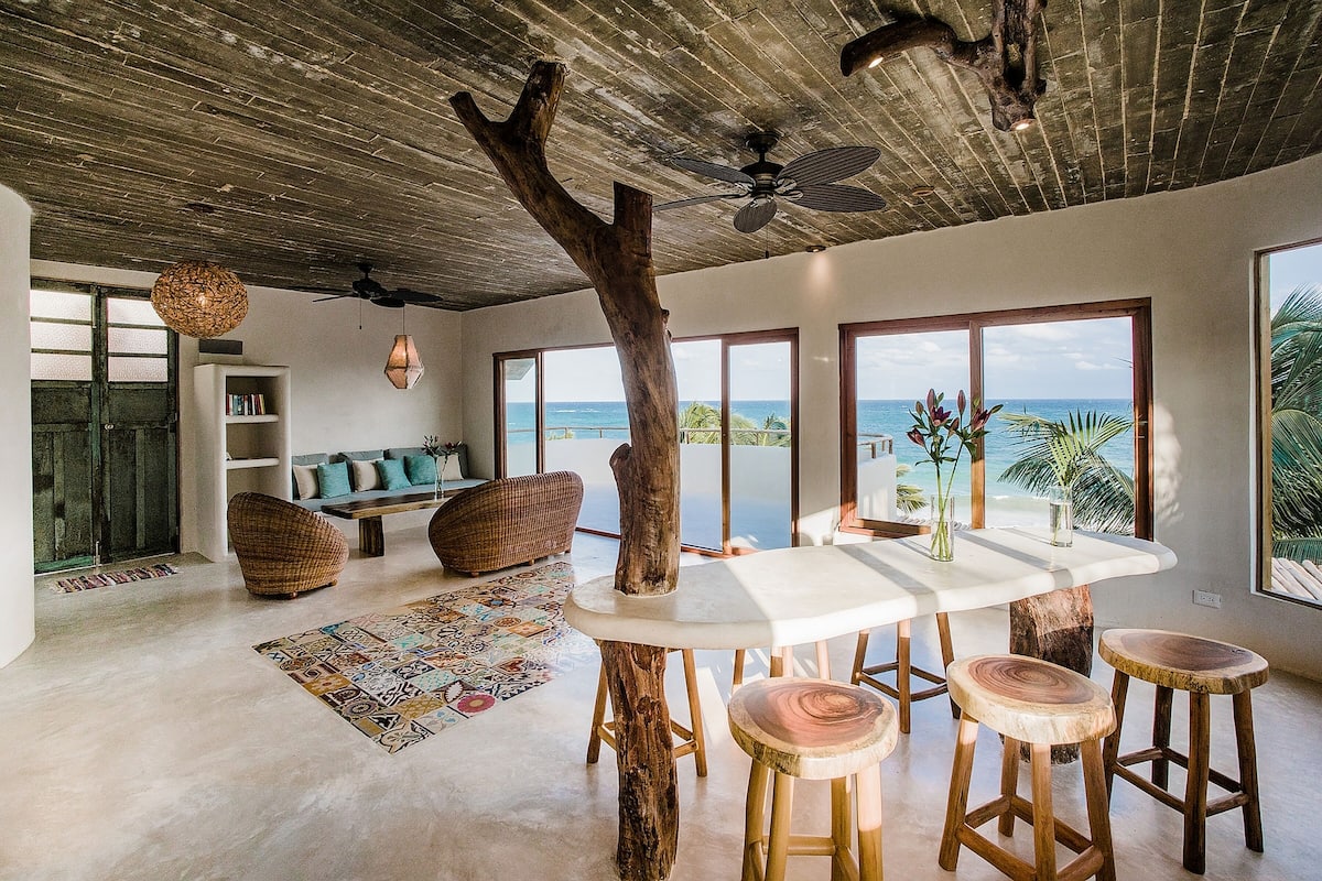 A spacious room with a large wooden table, wooden stools, a ceiling fan, and a view of the ocean.