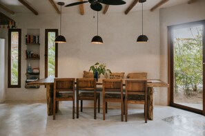 Un comedor con mesa y sillas de madera, luces colgantes y vistas a la vegetación a través de un ventanal grande.