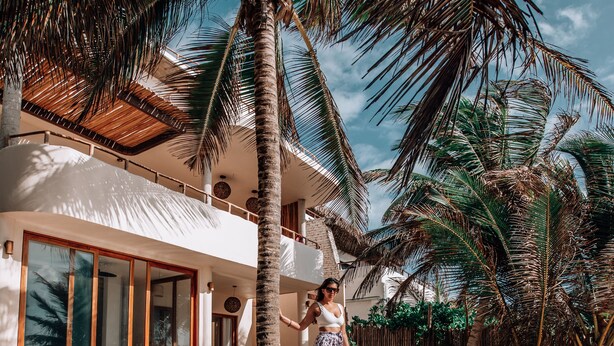 A poolside area with a palm tree, a pool, and a building with large windows and a balcony. - Zorba Tulum Beach Homes (Tulum)