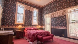 A bedroom with floral wallpaper, a bed with a red bedspread, two bedside tables with lamps, and a window with lace curtains. - Entire Historical Bed and Breakfast in Rico, Cololorado (Rico)