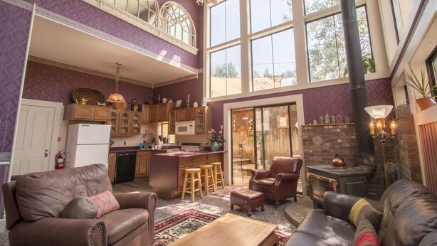 A spacious living area with a high ceiling, a fireplace, and a kitchen with wooden cabinets and a large window. - Entire Historical Bed and Breakfast in Rico, Cololorado (Rico)