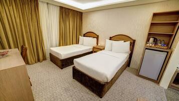A hotel room with two single beds, a small desk, a refrigerator, and a shelf with toiletries.