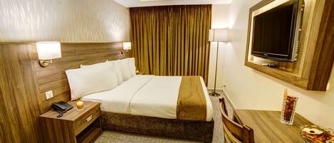 A hotel room with a bed, bedside table with a phone, a desk with a TV, and a chair.