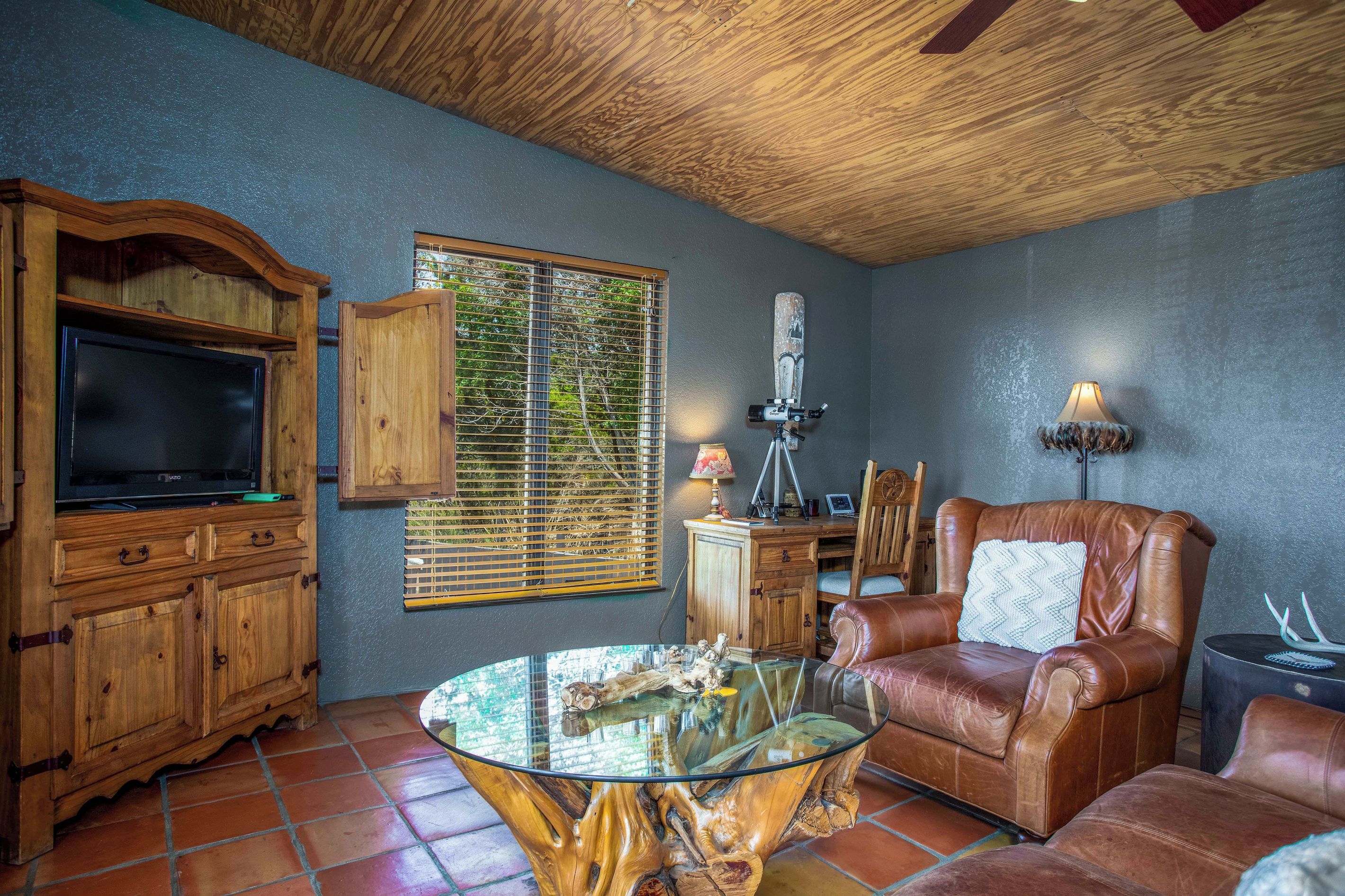 Una sala de estar con televisor, armarios de madera, una mesa de centro de vidrio, sofá de cuero y una ventana con persianas.