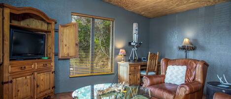 Una sala de estar con televisor, armarios de madera, una mesa de centro de vidrio, sofá de cuero y una ventana con persianas.