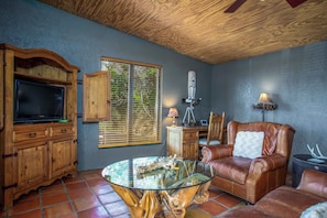 A living room with a television, wooden cabinets, a glass coffee table, a leather sofa, and a window with blinds. - Pipe Creek Ranch | Pets ok | Hot Tub | Sleeps 3 (Pipe Creek)