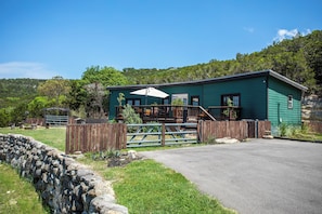 A green building with a porch, a stone wall, and a fenced area. - Pipe Creek Ranch | Pets ok | Hot Tub | Sleeps 3 (Pipe Creek)