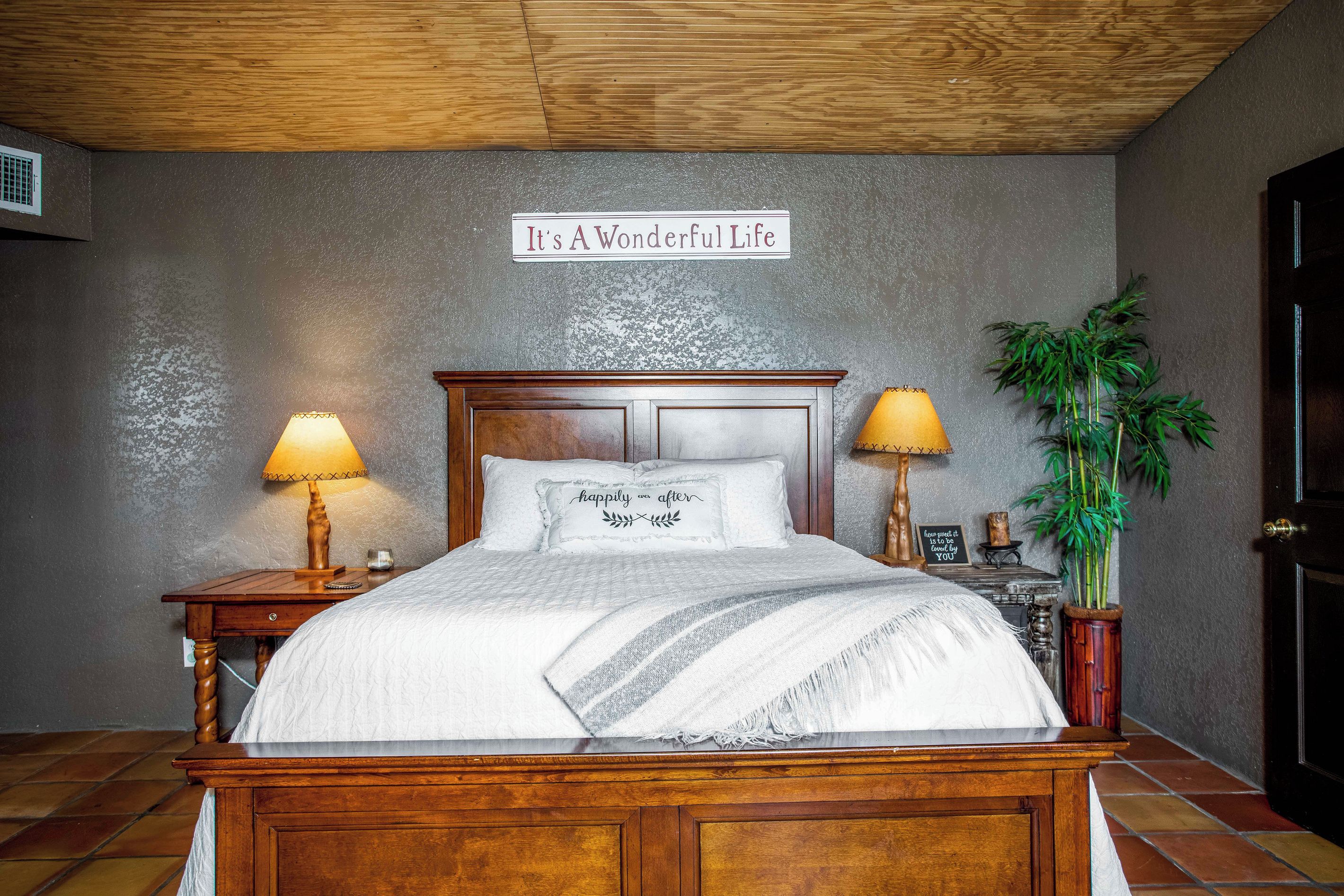 A bedroom with a wooden bed, two bedside lamps, a sign above the bed, and a potted plant.