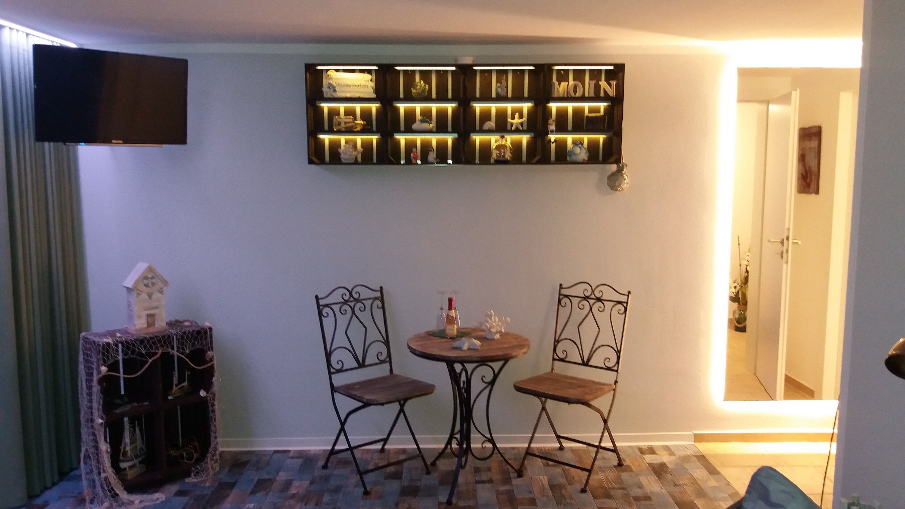 A room with a flat-screen TV mounted on the wall, a small table with two chairs, and a decorative shelf with various items.