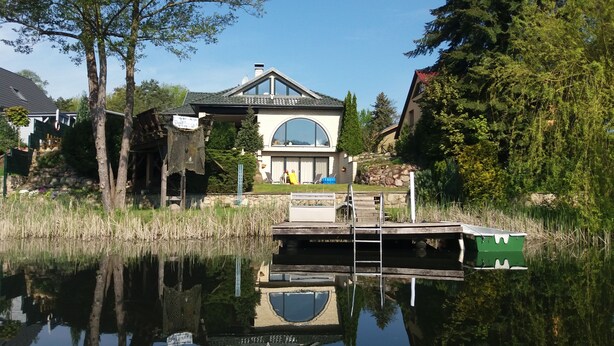 Une maison avec un quai s’avançant dans un lac paisible, entourée d’arbres et sous un ciel dégagé.